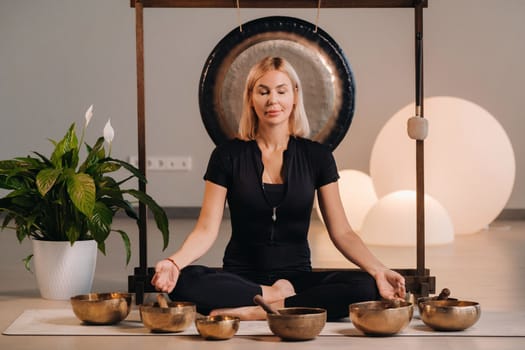 A woman sits in a lotus position next to Tibetan bowls, sitting on a yoga mat against the background of a gong.