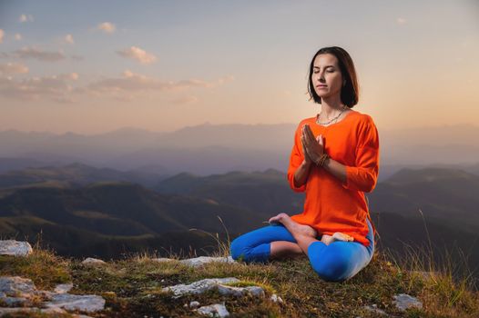 A young woman practices yoga outdoors in a beautiful mountain landscape. Against the background of sunsets, lotus position and namaste. Meditation and relaxation.