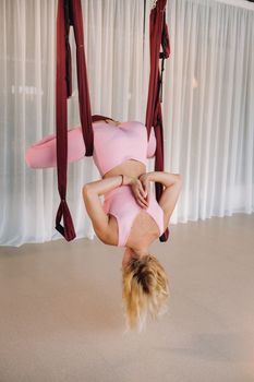 A young girl, an aero yoga coach, is hanging on a hammock in the yoga club hall. a woman in pink sportswear is doing anti-gravity yoga.