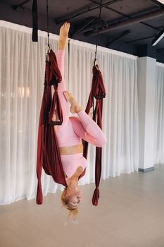 A young girl, an aero yoga coach, is hanging on a hammock in the yoga club hall. a woman in pink sportswear is doing anti-gravity yoga.