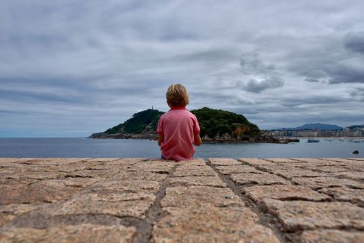 Blond boy on his back looking at the sea. Caucasian, San Sebastian, sky with clouds, mountain, textures, not identifiable