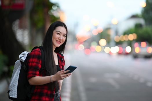 Young asian woman with backpack calling taxi on smart phone and standing on street.