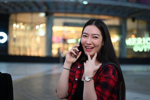 Young Asian woman talking on mobile phone while standing at outdoor shopping mall in the evening.