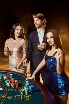 Two beautiful women and young man play on poker table in casino, focus on man and brunette. Passion, cards, chips, alcohol, dice, gambling, casino - it is entertainment. Dangerous fun card game for money. Smoke background.