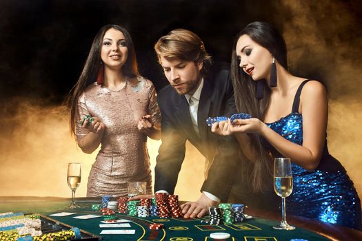 Two beautiful women and young man play on poker table in casino, focus on man and brunette. Passion, cards, chips, alcohol, dice, gambling, casino - it is entertainment. Dangerous fun card game for money. Smoke background.