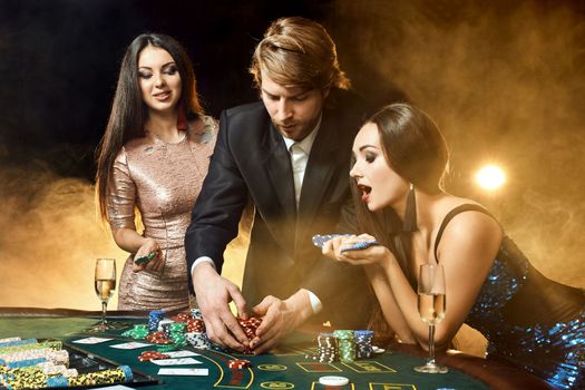 Two beautiful women and young man play on poker table in casino, focus on man and brunette. Passion, cards, chips, alcohol, dice, gambling, casino - it is entertainment. Dangerous fun card game for money. Smoke background.