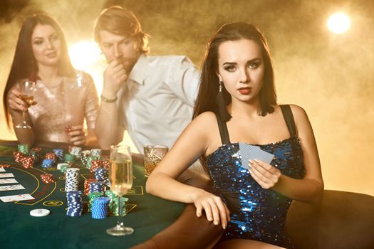 Group of an elegant people playing poker at the gambling house. Focus on a emotional brunette in a blue shiny dress. Passion, cards, chips, alcohol, dice, gambling, casino - it is entertainment. Dangerous fun card game for money. Smoke background.