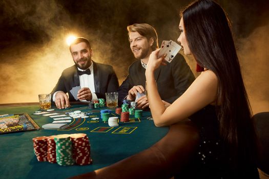Group of young rich people is playing poker in the casino. Two men in business suits and a young woman in a black dress. Smoke. Casino. Poker
