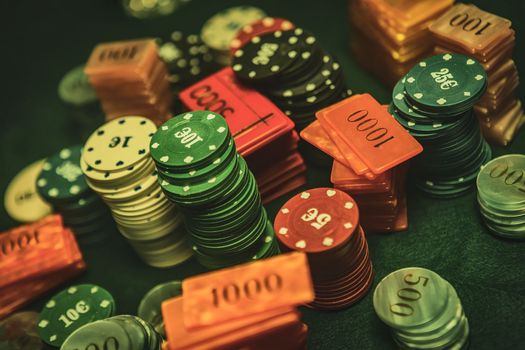 Poker chips are stacked neatly on a green textured table, the focus is on the chips with a shallow depth of field. The background is hazy with the smoke of cigars filling the room.