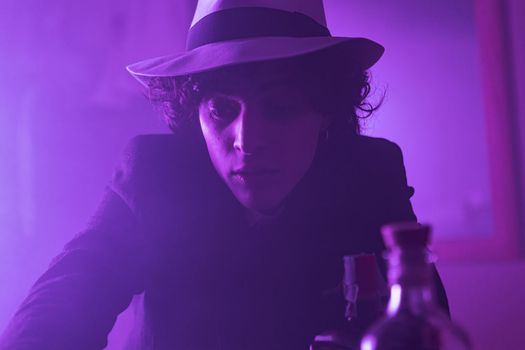 A young man with curly, brunette hair is looking at the camera with a smile. He is wearing a hat and suit. He appears confident and poised. The dark background adds a sense of mystery and intrigue.