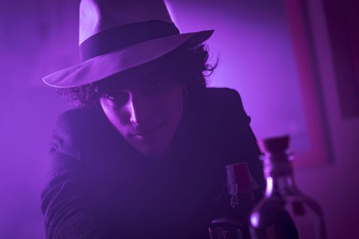 A young man with curly, brunette hair is looking at the camera with a smile. He is wearing a hat and suit. He appears confident and poised. The dark background adds a sense of mystery and intrigue.