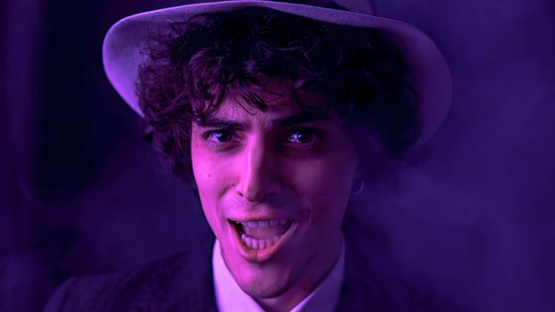 A young man with dark brown hair is happily singing and dancing with comical facial expressions in front of a smoky, dark background. He is wearing a stylish black suit and a fashionable hat.