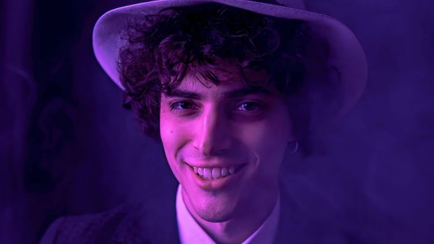 A young man with dark brown hair is happily singing and dancing with comical facial expressions in front of a smoky, dark background. He is wearing a stylish black suit and a fashionable hat.