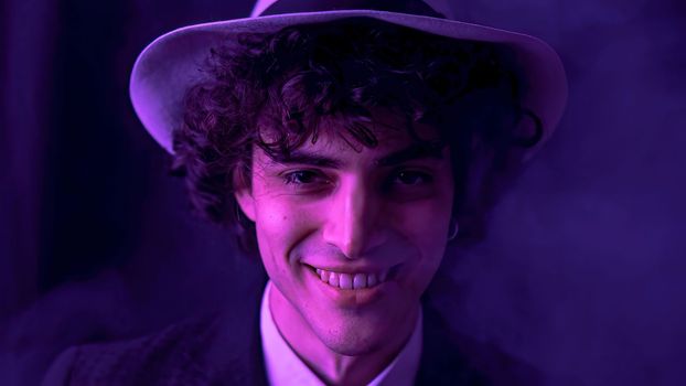 A young man with dark brown hair is happily singing and dancing with comical facial expressions in front of a smoky, dark background. He is wearing a stylish black suit and a fashionable hat.