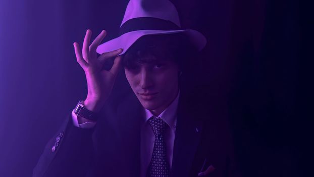 A handsome, curly haired man strikes a playful pose as he dances in a sleek black suit. Touching his a cowboy hat and a mischievous wink towards the camera. Against a dramatic, dark background.