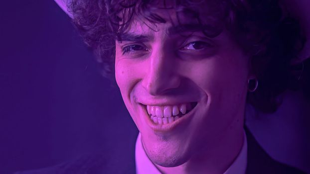An attractive young man with messy hair and a chiseled jawline beams at the camera with a toothy smile. With a background of deep darkness, this handsome individual exudes charm and inviting expression.
