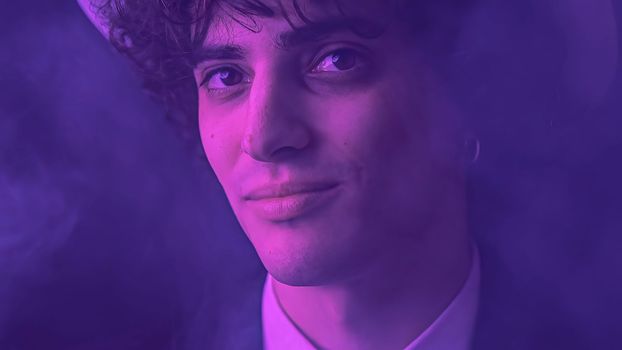 An attractive young man with messy hair and a chiseled jawline beams at the camera with a toothy smile. With a background of deep darkness, this handsome individual exudes charm and inviting expression.