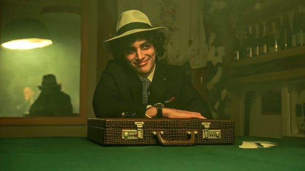 A man sitting at a table in a dimly lit room filled with smoke. He accepts a briefcase with a smile on his face. The scene suggests a negotiation or possibly a shady deal.