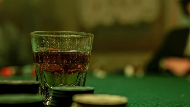A man is pouring whiskey into a glass placed on a poker table covered in green felt. Surrounding the glass are stacks of poker chips, hinting at a game in progress or about to begin.