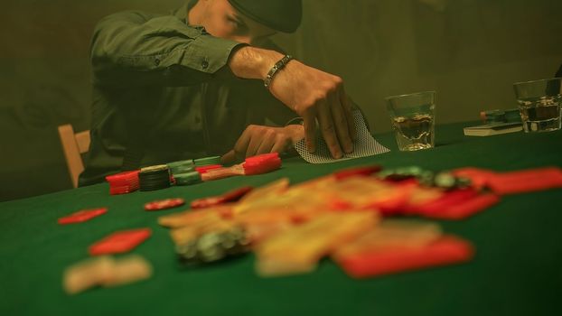 A drunk poker player sipping whiskey while checking his cards placed in front of him on the green textured table. He adds all his poker chips to the pile on the table signalling an all-in bet.