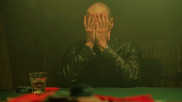 A worried poker player sits at a dimly lit table, nervously sipping on a glass of whiskey. The player pushes all chips forward, signaling an all-in bet, raising the stakes.