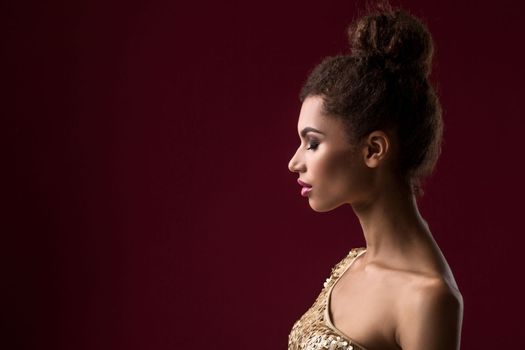 Fashion young African woman with make-up, in sexy gold dress. Model on a claret background in the studio. A woman is half a turn