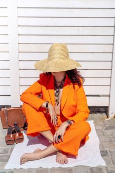 Stylish woman in an orange suit with a hat sits on a rug on a white striped background. On the hands are jewelry rings and bracelets, sandals and a bag stand side by side