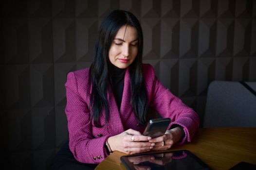 Beautiful confident Caucasian 40-45 years old woman, successful businesswoman in formal wear, chatting on mobile phone while waiting for colleagues and business partners for meeting in office interior