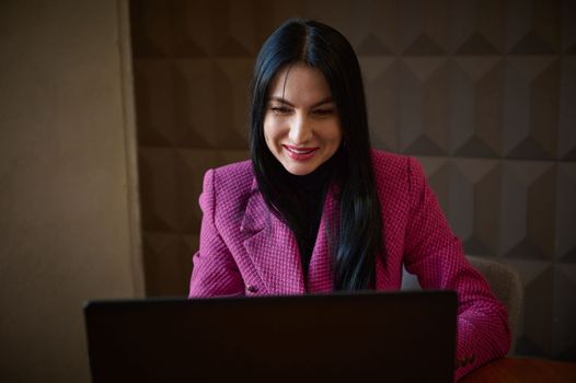 Smiling confident woman entrepreneur 40-45 years old, dressed in a stylish crimson jacket, talking to business clients via video link. Successful entrepreneur at online meeting with business partners
