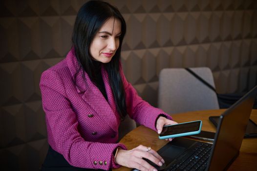 Successful confident 40-45 years old middle-aged Caucasian woman, entrepreneur in casual wear, holding mobile phone and typing text on laptop in a cozy office interior. People, online business concept