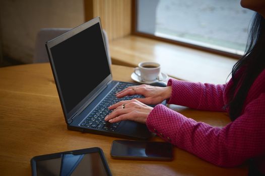 Selective focus on hands of a businesswoman, dressed in bright magenta jacket, on laptop keyboard while telecommuting, sitting at table with a cup of coffee in a cozy cafeteria. Remote work. Business