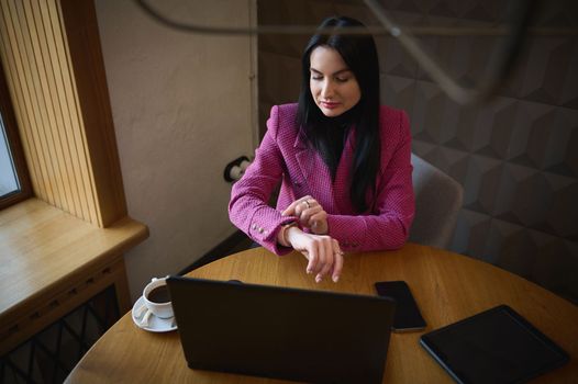 Top view Caucasian successful purposeful business woman, looking at wristwatch, tracking time on smartwatch, sitting at table with laptop and waiting for meeting with investors in an office interior.