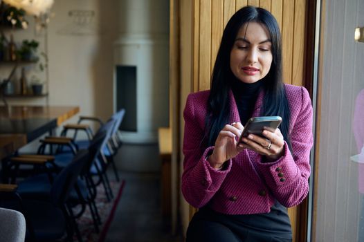 Confident elegant dark-haired Caucasian pretty woman 40s, sitting on the window sill in a meeting room, chatting on her modern smartphone, planning meeting with business partners, investors, employees