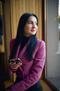 Beautiful middle-aged 40s woman in stylish wear, holding smartphone and thoughtfully looking at the urban background through the window, sitting on the windowsill in a cozy interior of a coffee house
