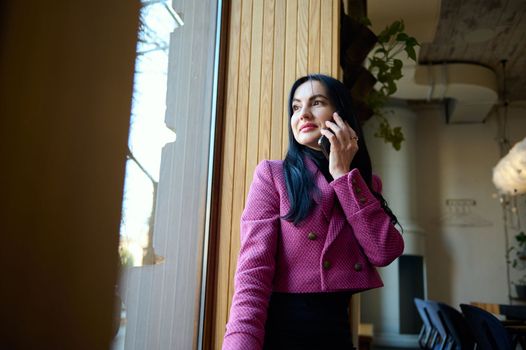 Confident middle-aged Caucasian woman in stylish formal wear, sitting on the window sill, talking on mobile phone, looking through the window in conference room. People. Communication. Business