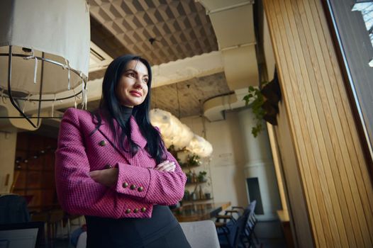 Portrait of beautiful goal-oriented businesswoman, restaurateur, executive director, standing with her arms crossed in creative art cafe and looking aside while smiling. Successful people concept