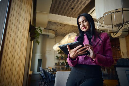 Modern stylish middle-aged successful woman entrepreneur, sales manager, chief executive working remotely on a digital tablet from the interior of a restaurant. People and business. Low angle view