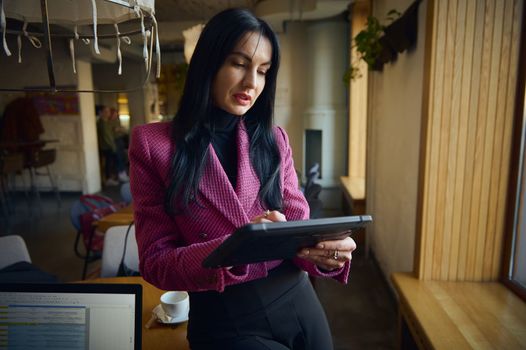 Middle-aged charming businesswoman holding in hands digital tablet while standing in modern restaurant interior, successful female CEO dressed in elegant clothes posing while work on portable net-book