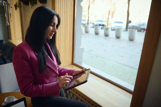 Confident elegant stylish middle aged Caucasian business woman, entrepreneur, chief-executive standing by window sill, working on digital tablet, writing weekly report during coffee break in a cafe
