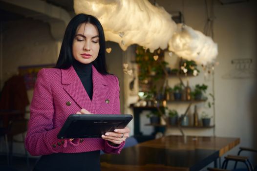 Charming middle-aged dark-haired Caucasian woman in elegant stylish casual wear, purposeful restaurateur, CEO, successful manager, business lady working on tablet in the modern restaurant interior