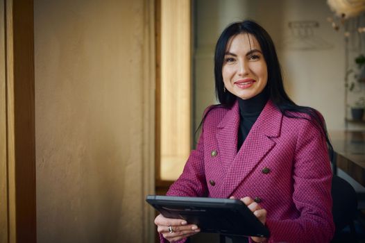 Smiling middle-aged Caucasian business woman, leader entrepreneur, CEO, professional manager holding digital tablet computer, using software applications standing in the interior of modern restaurant