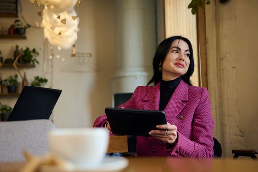 Middle-aged Caucasian confident, successful gorgeous woman, wearing stylish purple casual jacket, working on a digital tablet, waiting for business partners while a coffee break in a cozy restaurant