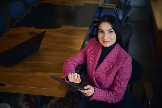 Attractive purposeful middle aged businesswoman, CEO, holding digital tablet, smiling cutely looking at camera, looking forward to meeting with business partners and investors at the negotiation table