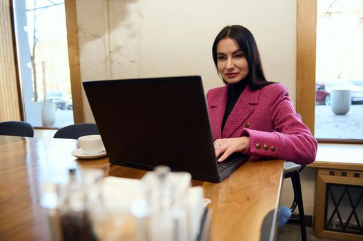 Confident Caucasian woman entrepreneur in purple casual jacket, working remotely on laptop in a coffee shop interior. Successful business lady telecommuting, planning new actions for her business plan