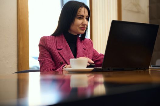 Successful Caucasian middle-aged woman entrepreneur, business person in casual wear, sitting at table with laptop and cup of coffee, while an online meeting with business partners, in office interior