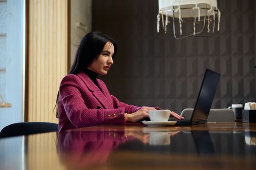 Confident successful woman entrepreneur, developing an action plan for her future business project, sitting at table with a cup of hot coffee drink and working on laptop in a VIP lounge zone
