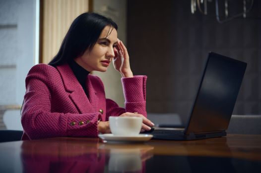 Confident Caucasian middle-aged business woman, entrepreneur, working remotely on laptop with a cup of coffee in cozy interior, planning new project, online chatting with business partners and clients