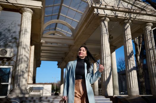 Beautiful dark-haired Caucasian middle-aged woman in casual wear, cheerfully smiling at camera while walking down the urban street on a beautiful sunny early spring day. People. Urban. life Lifestyle