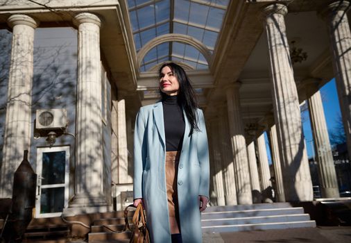 Caucasian elegant stylish dark-haired middle aged pretty woman in stylish blue coat, holding handbag, enjoying morning walk on the city street on a sunny early spring day. People and lifestyle concept