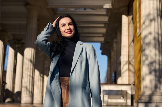 Charming stylish middle-aged Ccaucasian woman, pretty brunette in elegant blue warm coat, strolling along the city street on a sunny day, fixing her hairstyle while the wind is blowing her long hair